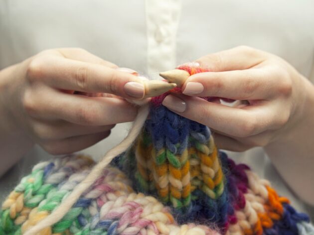 Young woman knitting a woolen colorful scarf. She is using bamboo knitting needles, size ten. Only her hands and arms are showing on the image. She has manicured nails. Neutral colored nailpolish. She is wearing an ivory colored blouse with transparant buttons. Rolled up sleeves, bare arms are visible. The knit has pastel colors as well as brighter colors. Pink, blue, green, orange, ecru and yellow.
